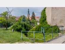 Eine kleine Grünfläche mit einem schmalen Bachlauf, umgeben von Bäumen und Büschen. Im Hintergrund sind Dächer von Häusern und ein Kirchturm mit einer Turmspitze zu sehen, rechts im Bild eine mit Pflanzen bewachsene Hauswand.