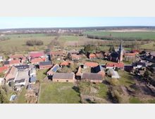 Luftaufnahme eines Dorfes mit verstreuten Wohnhäusern und landwirtschaftlichen Gebäuden aus Ziegeln und Fachwerk, umgeben von Wiesen und Feldern. Im Mittelpunkt steht eine Kirche mit hohem Turm und spitzem Dach, daneben führen Straßen und Baumreihen durch die ländliche Umgebung.