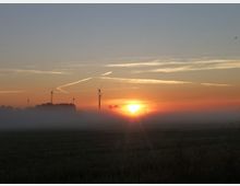 Eine Landschaft bei Sonnenaufgang mit leichtem Nebel, im Vordergrund ein Feld, im Hintergrund mehrere Windräder und Baumgruppen, der Himmel ist in Orange- und Blautönen gefärbt.