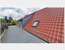 Flachdach mit angrenzendem, geneigten Dach mit roten Dachziegeln und einem Dachfenster; umgeben von Bäumen und einigen Hausdächern im Hintergrund.