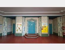 Innenraum eines gefliesten Treppenhaus- oder Flurbereichs mit rotbraunem Boden und mehreren türkisfarbenen Türen, die von blau gemusterten Fliesenbändern gerahmt sind. In der Mitte führt eine kurze, gestufte Plattform zu einer zentralen Tür; an den Wänden hängen farbige Plakate und rechts sind Feuerlöscher angebracht.