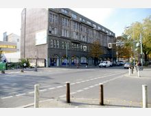 Kreuzung in einer städtischen Umgebung mit einem vierstöckigen Gebäude aus dunklem, älterem Stein und großen Fenstern im Hintergrund. Im Vordergrund befinden sich ein Fußgängerüberweg, parkende Autos, Radfahrer und Fußgänger sowie einige Bäume am Straßenrand.