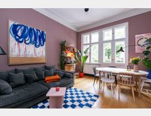 Ein gemütliches Wohnzimmer mit pastellvioletten Wänden, einem großen grauen Sofa, dekoriert mit Kissen, und einem kleinen rosa Beistelltisch auf einem blau-weiß karierten Teppich. Im Hintergrund befindet sich ein heller Essbereich mit einem ovalen Holztisch, verschiedenen Stühlen und großen Fenstern, die Tageslicht hereinlassen; Pflanzen und farbenfrohe Kunstwerke ergänzen den Raum.