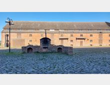 Historisches Hofgelände mit gepflastertem Boden, zentraler Backsteinstruktur und einem eingebauten Ofen oder Grill. Im Hintergrund steht ein großes, langgestrecktes Backsteingebäude mit Holztüren und kleinen Fenstern unter einem Spitzdach.