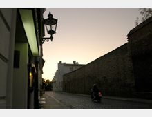 Eine gepflasterte Straße in einer urbanen Umgebung bei Sonnenuntergang, flankiert von einer hohen Backsteinmauer auf der rechten Seite und beleuchteten Gebäuden links. Ein Motorradfahrer ist auf der Straße zu sehen, während eine historische Straßenlaterne im Vordergrund hängt.