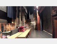 Blick hinter die Bühne eines Theaters mit sichtbaren Bühnentechnik-Elementen wie Seilzügen, Vorhängen und einer metallenen Tür im Hintergrund. Im Vordergrund stehen gestapelte Stühle, Tische und Sitzpolster.