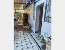 Veranda mit weiß gefliesten Bodenfliesen und dekorativen Glasfenstern, teilweise überdacht mit einer durchsichtigen Dacheindeckung. Im Vordergrund stehen ein Rattanstuhl und Pflanzen, während im Hintergrund ein Essbereich mit Stühlen und ein Zugang zu einem Garten sichtbar sind.