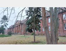 Großes Gebäude aus rotem Backstein mit mehreren Fenstern und einem mit Säulen versehenen Eingangsbereich. Im Vordergrund befindet sich eine Wiese mit kahlen Bäumen und einem Nadelbaum, während der Himmel bedeckt ist.