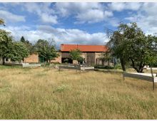 Eine ländliche Szene mit einer Wiese, mehreren Obstbäumen und einigen erhöhten Beeten aus Holz. Im Hintergrund ist ein Gebäude aus Backstein mit einem roten Ziegeldach und großen Holztoren zu sehen.