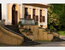 Gelbes historisches Gebäude mit verzierten dunklen Fenstern und offenen Fensterläden. Eine symmetrische Treppe mit geschwungenen Geländern führt zu einem erhöhten Balkonbereich, der mit Blumenkübeln dekoriert ist; im Hintergrund ist üppiges Grün sichtbar.