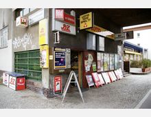 Kiosk in einer städtischen Umgebung mit mehreren Werbeschildern, Zeitungsständern und Lottoangeboten. Die Fassade ist teilweise mit Graffiti bedeckt, und der Eingangsbereich zeigt Zeitungen, Plakate und Aushänge vor einem kleinen Laden.