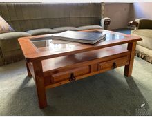 Wohnzimmer mit grün gepolsterter Couch und Sessel auf Teppichboden, davor ein rechteckiger Holzcouchtisch mit Glasplatte und zwei Schubladen. Auf dem Tisch liegt ein großes Buch, im Hintergrund ist ein Fensterbereich zu sehen.