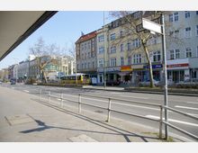 Straßenszene in einer Stadt mit mehrstöckigen Gebäuden, darunter Geschäfte und Cafés im Erdgeschoss. Am Straßenrand sind parkende Autos, ein Linienbus und einige Bäume zu sehen; ein Straßenschild im Vordergrund weist auf die "Treitschkestraße" hin.