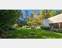 Großer Garten mit gepflegtem Rasen und verschiedenen Bäumen und Sträuchern, umgeben von dichter Vegetation. Rechts im Bild ein modernes Gebäude mit weißer Fassade und großen Fenstern.