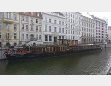 Ein historisches Schiff mit der Aufschrift „HANSEAT“ liegt an einem Kanal vor einer Reihe von mehrstöckigen, hellen Wohn- und Bürogebäuden im traditionellen Stil. Die Architektur der Gebäude zeichnet sich durch weiße Fassaden und Sprossenfenster aus.