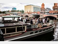 Ein Boot mit einer Menschenmenge, die offensichtlich an einer Feier teilnimmt, liegt vor Anker auf einem Fluss. Im Hintergrund ist die Oberbaumbrücke in Berlin zu sehen, zusammen mit umliegenden Gebäuden und einer urbanen Szenerie.