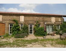 Backsteinfassade eines rustikalen Gebäudes mit großen Bogenfenstern und einer Schiebetür aus Holz. Davor befindet sich ein kleiner Gartenbereich mit einem Sonnenschirm, Holzmöbeln und dichter Begrünung.