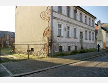 Straßenansicht eines alten, heruntergekommenen Gebäudes mit sichtbaren Putzschäden und freiliegendem Mauerwerk. Im Vordergrund ein offenes Metalltor, Kopfsteinpflaster und eine einzelne Gehperson; im Hintergrund ein weiteres Gebäude mit roten Ziegeln.