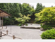 Japanischer Steingarten mit sorgfältig geharktem Kies, umgeben von grünen Bäumen und Sträuchern. Links im Vordergrund befindet sich eine Holzüberdachung mit einer dekorativen Metallkette, die als Regenrinne dient.