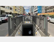 Eingang zu einer U-Bahn-Station mit Treppen, umgeben von einem Metallgeländer. Im Hintergrund eine belebte Straßenansicht mit geparkten Autos, Gebäuden und Gehwegen auf beiden Seiten.