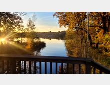 Holzgeländer mit Blick auf einen ruhigen See, umgeben von herbstlichen Bäumen mit gelben und orangefarbenen Blättern. Im Hintergrund sind Häuser am Ufer unter einem klaren Himmel zu erkennen, während die tiefstehende Sonne durch die Bäume scheint.