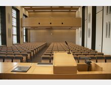 Ein moderner Hörsaal mit Holzelementen, Reihen von gepolsterten Sitzen, einem Rednerpult mit Mikrofon im Vordergrund und großen Fenstern an der linken Wand. Die Rückwand besteht aus Holzpaneelen, und eine analoge Wanduhr hängt zentral an der Wand.