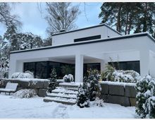 Modernes, weißes Flachdachhaus mit großen Glasfronten und überdachter Terrasse, davor eine niedrige Natursteinmauer mit Treppe. Der verschneite Garten mit Sträuchern und Bäumen umgibt das Gebäude in winterlicher Umgebung.