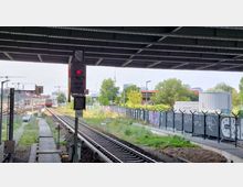 Blick auf ein Bahngleis unter einer Brücke mit einem roten Signal im Vordergrund. Im Hintergrund sind ein S-Bahn-Zug, Gebäude, der Berliner Fernsehturm und ein Zaun mit Graffiti sichtbar.