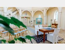 Heller Innenraum einer kleinen Kirche mit hellen Wänden und gotischen Bögen. Im Vordergrund steht ein Holztisch mit weiß überzogenen Stühlen, im Hintergrund sind Holzbänke und eine kleine Orgel sichtbar.