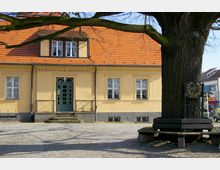 Gelbes Gebäude mit rotem Ziegeldach, beschriftet mit "Heimatmuseum Zehlendorf", und einer zentralen grünen Eingangstür. Im Vordergrund ein großer Baum mit einem umlaufenden Holzsitzbank und einem dekorativen Schild.