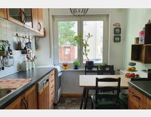 Kleine Küche mit Holzmöbeln und schwarzer Arbeitsplatte, einem Esstisch mit Stühlen und einem großen Fenster mit Blick auf einen Hinterhof mit Bäumen. Auf der Fensterbank stehen Zimmerpflanzen, und an der Wand hängt ein Regal mit Küchenutensilien.