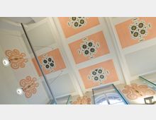 Decke eines historischen Gebäudes mit dekorativen, geometrischen Mustern in Pastelltönen, insbesondere rosa und weiß, sowie symmetrischen, kunstvollen Verzierungen. Im unteren Bereich sind Fenster mit Rundbögen und Glasscheiben erkennbar, ergänzt durch moderne Lampen.