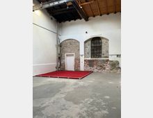 Innenraum einer ehemaligen Industriehalle mit hohen weißen Wänden und freigelegtem Ziegelmauerwerk in Rundbogen-Nischen, darunter ein kleines weißes Türportal. Rechts befindet sich ein großes, bogenförmiges Sprossenfenster; die Decke besteht aus Holz und sichtbaren Stahlträgern, am Boden steht eine niedrige Bühne mit rotem Teppich.