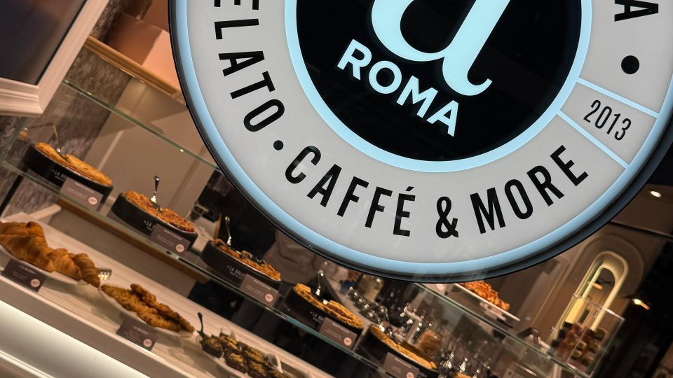 Ein modernes Café namens "La Dolceria" mit einem beleuchteten Rundlogo, das "Gelato, Caffè & More" zeigt. Im Vordergrund eine Vitrine mit einer Auswahl an Gebäck, Kuchen und anderen süßen Speisen.