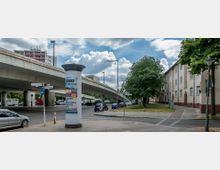 Straßenszene in einer urbanen Umgebung mit einer Hochstraße im Hintergrund, Wohngebäuden rechts und einem begrünten Gehweg. Im Vordergrund steht eine Litfaßsäule mit Plakaten, daneben parkende Autos und Verkehrsschilder.