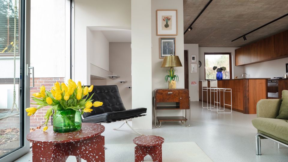 Open-plan modern living room and kitchen with a raw concrete ceiling, large floor-to-ceiling sliding glass doors, and white floors. The space includes a black lounge chair, green sofa, terrazzo-style red tables on a light rug, and a wood-fronted kitchen with bar stools along the counter.