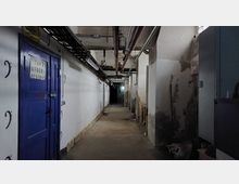Ein schmaler, karger Korridor in einem Keller, mit weißen, teilweise abgenutzten Wänden, sichtbaren Rohren und Kabeln an der Decke, einer blauen Metalltür links und einigen aufgestapelten Gegenständen rechts. Der Gang ist schwach beleuchtet und führt ins Dunkel.