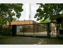 Eingangstor zu einem umzäunten Gelände mit gepflastertem Weg und einem Backsteingebäude im Hintergrund. Im Zentrum ragt ein hoher, weißer Turm empor, umgeben von Bäumen.