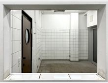 Blick durch ein rechteckiges Fenster auf einen gefliesten, leeren Raum mit weißen Wandfliesen und grauen Bodenfliesen. Links ist eine Holztür mit rundem Fenster, rechts eine graue Tür, und an der Decke fehlen teilweise Platten.