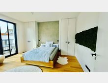 Schlafzimmer mit einem Doppelbett auf einem Holzrahmen, weißen Wänden und einem grauen Paneel an der Rückwand. Links führt eine Glasschiebetür zu einem Balkon, rechts ist eine Mooswand dekorativ angebracht und der Boden besteht aus hellem Holzparkett.