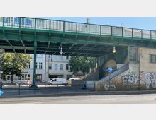 Grüne Hochbahnbrücke über einer Straße mit Treppenaufgang zur U-Bahn-Station "Eberswalder Straße", daneben eingelassene Bögen im Mauerwerk und Graffiti an den Wänden. Im Hintergrund Geschäftsfassaden und Bäume.