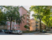 Mehrstöckiges Wohngebäude mit rosa Fassade, Balkonen und einem Logo der Baugenossenschaft auf der Seitenwand. Das Gebäude steht an einer von Bäumen gesäumten Straße mit parkenden Autos.