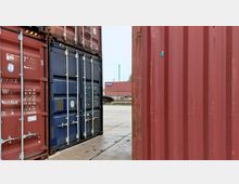 Mehrere übereinander gestapelte Schiffscontainer in Blau und Rot auf einem Betonplatz, mit Blick durch die Container auf weitere gestapelte Container und eine grüne Landschaft im Hintergrund. Ein Teil der Container zeigt Gebrauchsspuren und verblasste Aufschriften.