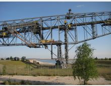 Eine große Stahlkonstruktion einer Abraumförderbrücke in einer Landschaft mit Wiesen, Bäumen, Sand und einem Gewässer im Hintergrund. Im Vordergrund steht ein einzelner Baum, und das Gebiet ist durch einen Metallzaun abgegrenzt.