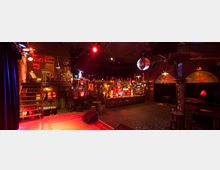 Ein gemütlicher Bar- und Veranstaltungsraum mit warmem, gedämpftem Licht, einer großen Theke im Hintergrund, dekoriert mit bunten Lampen und Flaschen, und einer erhöhten Bühne im Vordergrund. Eine Discokugel hängt von der Decke, und es gibt Sitzbereiche mit Polstermöbeln und Barhockern.