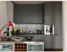 Moderne Küche mit grauen Einbauschränken, einer Rückwand im sechseckigen Fliesenmuster und Edelstahlgeräten, einschließlich eines eingebauten Backofens. Im Vordergrund liegt eine Theke mit Obstschalen, einem Weinregal und einer roten Schreibtischlampe.