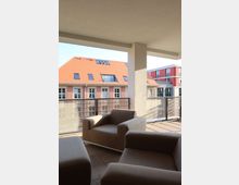 Überdachte Terrasse mit modernen beigen Sesseln und Ausblick auf ein Nachbargebäude mit ziegelrotem Dach und großen Fenstern. Im Hintergrund ist ein weiteres Gebäude mit roten und grauen Fassadenelementen zu erkennen.