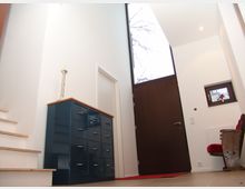 Moderner Eingangsbereich mit hohen weißen Wänden, einer dunklen Holztür mit einem großen, rechteckigen Fenster oben, einer kleinen Fensteröffnung rechts und einer Treppe links. Ein dunkelblauer Schrank mit Holzplatte und ein Stuhl mit rotem Polster runden das eingerichtetete Ambiente ab.