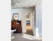 Minimalist interior room with a raw concrete ceiling, light gray walls, and carpeted floor, featuring a mid-century wooden cabinet and a white chair. An open white door leads to a hallway with built-in wood cabinetry and a small horizontal window, with sunlight casting striped shadows on the wall.