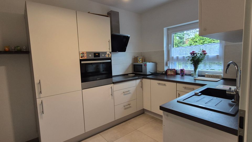 Moderne, helle Einbauküche in L-Form mit weißen Schränken, integriertem Backofen, schwarzer Arbeitsplatte und Edelstahlspüle. Großes Fenster über der Arbeitsfläche mit Blick ins Grüne sowie Deckenleuchte und Fliesenboden.